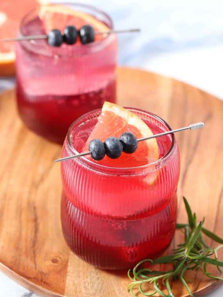 Glasses filled with purple drink, topped with grapefruit wedges and a skewer of blueberries.