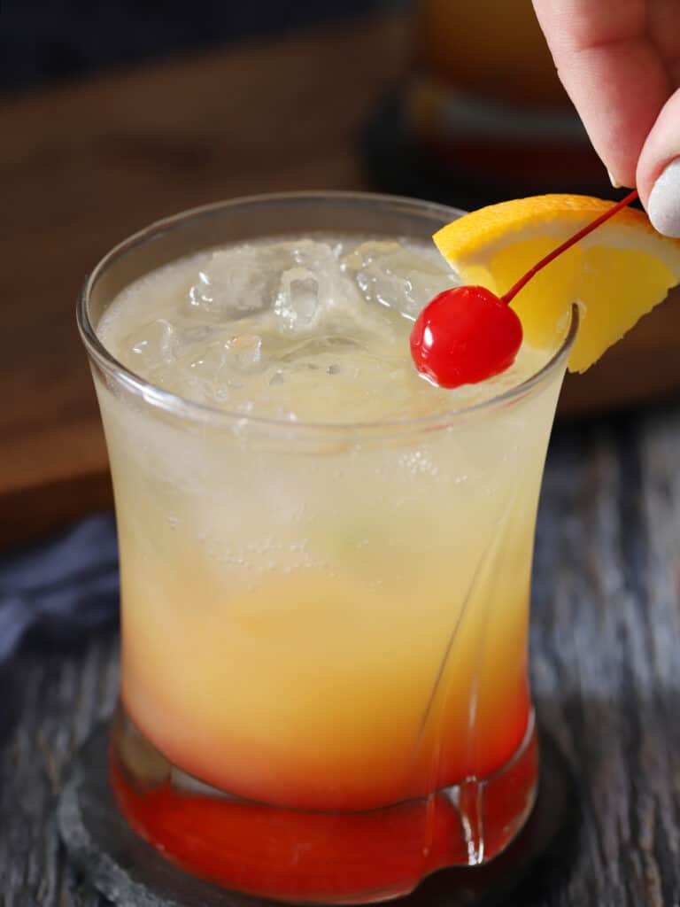 A hand placing cherry garnish on a layered drink in a glass.