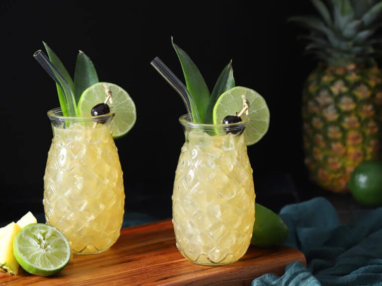 Pineapple shaped glasses filled with yellow cocktail, garnished with pineapple leaves, lime, and cherries.