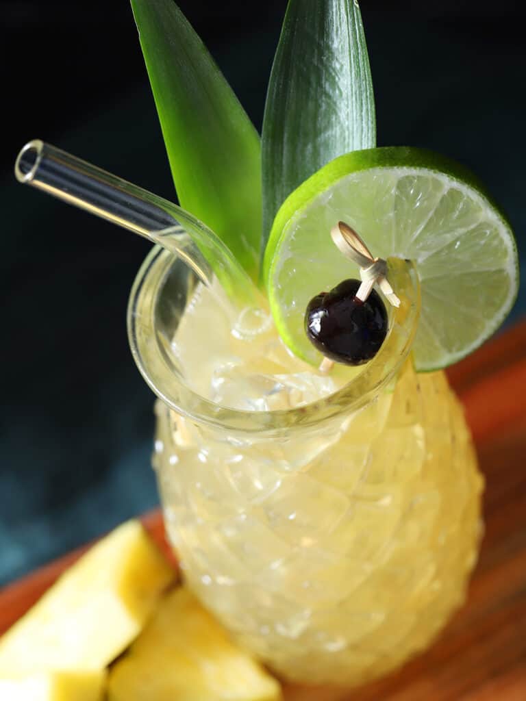 A close up of a pineapple glass filled with yellow cocktail and garnished with pineapple leaves, lime, and a cherry.