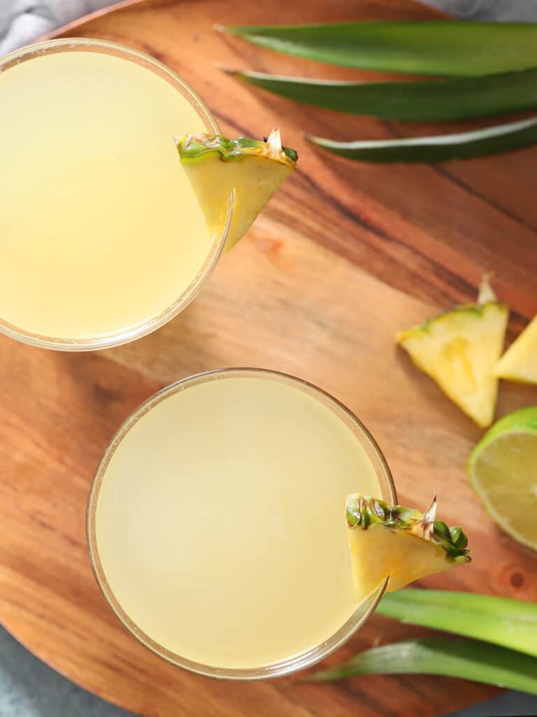Glass of yellow drinks on a tray with pineapple leaves and fresh pineapple pieces.