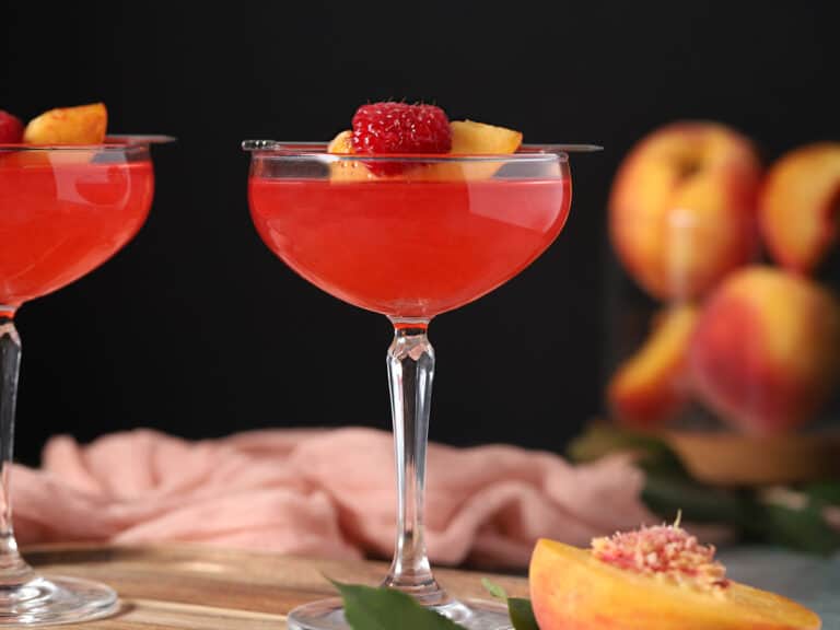 Glasses of raspberry martini garnished with peach slices and fresh raspberry.